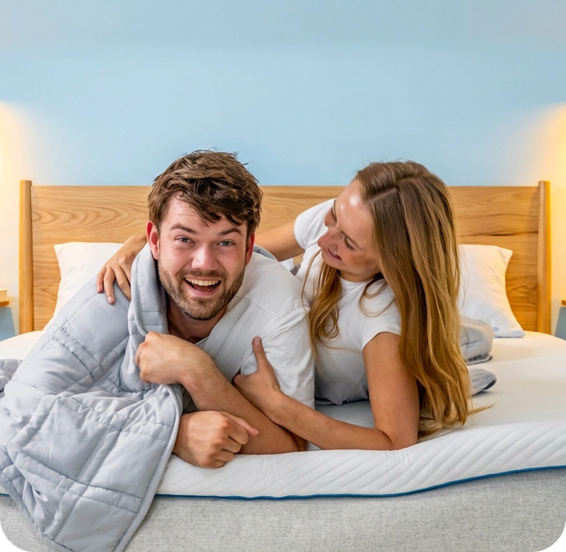 Lachend koppel ligt samen op bed onder een grijze Ella deken.